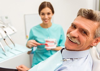 An older man smiling after receiving his customized restoration while a dental hygienist shows him how to properly care for his dental implants