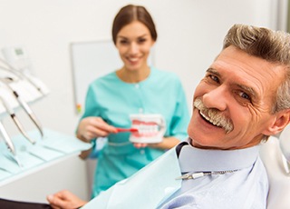 An older man smiling after receiving his customized restoration while a dental hygienist shows him how to properly care for his dental implants