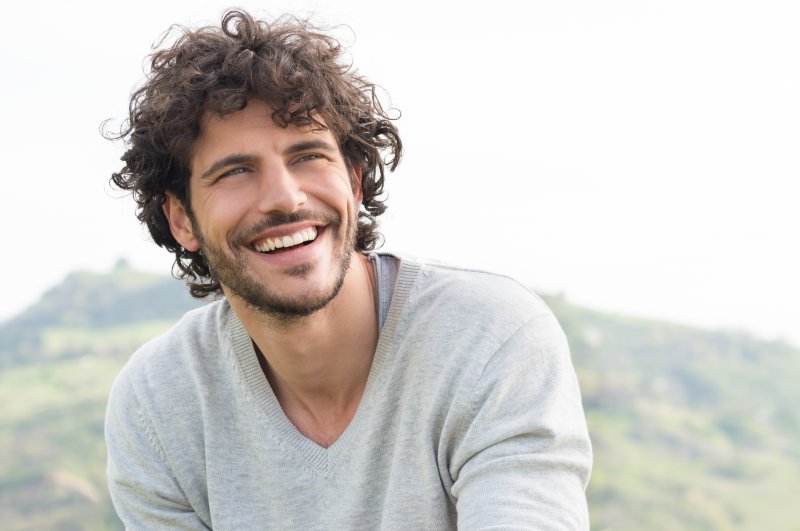 a young male wearing a gray V-neck sweater and sitting outside smiling